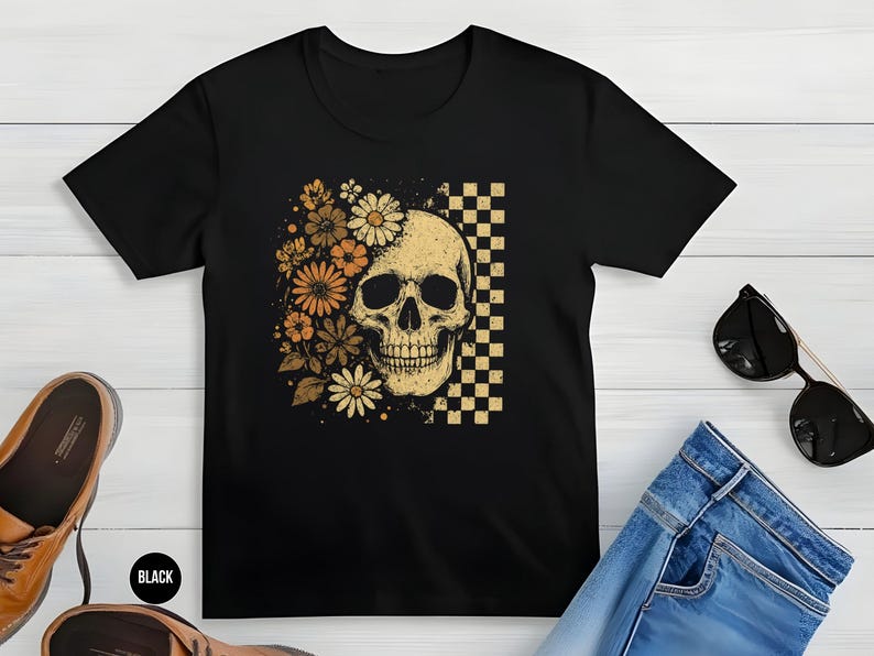 May include: Black t-shirt featuring a skull design with floral accents and a checkered pattern. The skull is cream-colored, and the flowers are in shades of orange and yellow. The shirt is paired with jeans and sunglasses.