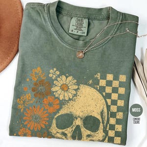 May include: Moss green t-shirt with a skull and floral design. The graphic features a skull, daisies, and a checkered pattern. A gold necklace with a pendant rests on the shirt. The shirt is labeled "Comfort Colors".