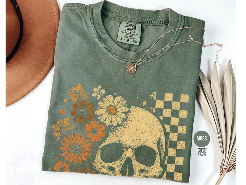 May include: Moss green t-shirt with a skull and floral design. The graphic features a skull, daisies, and a checkered pattern. A gold necklace with a pendant rests on the shirt. The shirt is labeled "Comfort Colors".