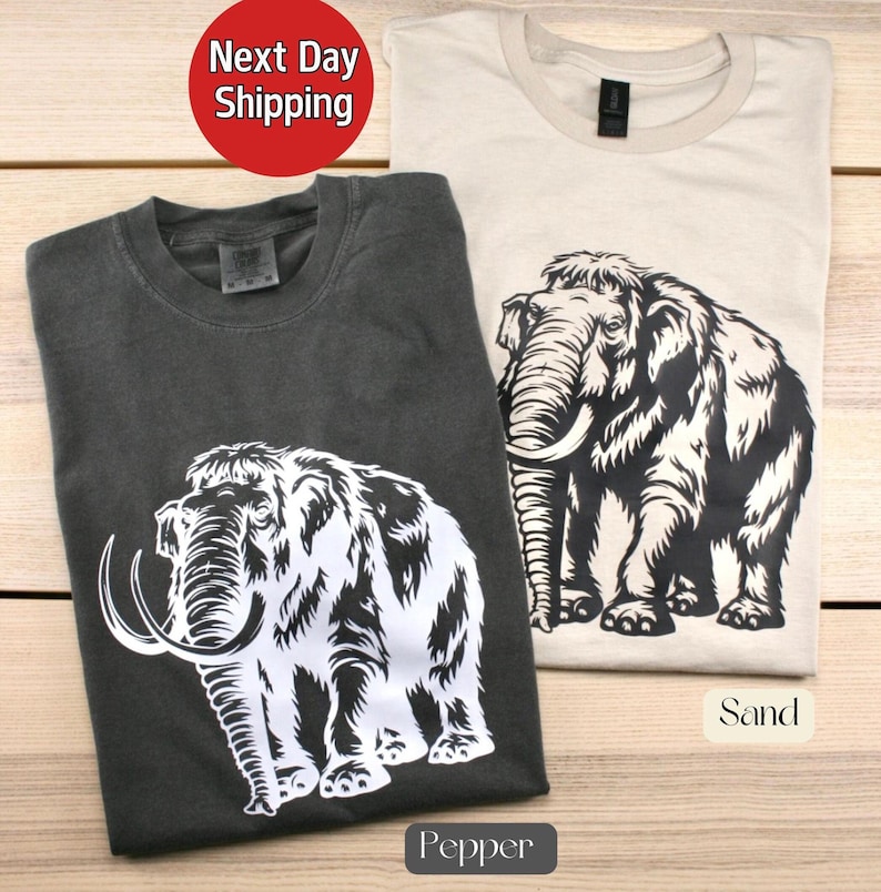 May include: Two t-shirts with a white woolly mammoth graphic. The shirt on the left is dark gray and says "Pepper" below the graphic. The shirt on the right is light beige and says "Sand" below the graphic.