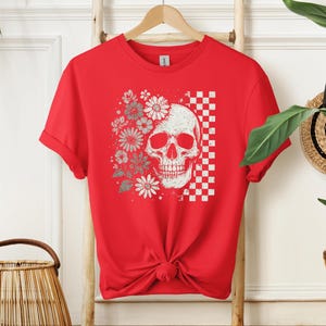 May include: Red t-shirt featuring a distressed skull graphic with floral and checkered elements. The design includes white and gray flowers and a checkered pattern. The shirt is hanging on a wooden ladder.
