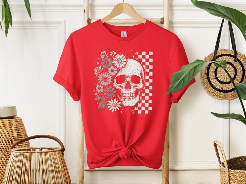 May include: Red t-shirt featuring a distressed skull graphic with floral and checkered elements. The design includes white and gray flowers and a checkered pattern. The shirt is hanging on a wooden ladder.