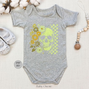 May include: A gray baby onesie with a neon green skull and floral design. The design includes yellow flowers and a checkered pattern. The onesie is surrounded by white baby shoes, cotton sprigs, and a wooden pacifier clip.