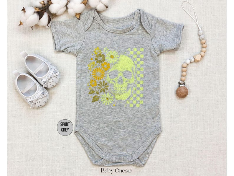 May include: A gray baby onesie with a neon green skull and floral design. The design includes yellow flowers and a checkered pattern. The onesie is surrounded by white baby shoes, cotton sprigs, and a wooden pacifier clip.