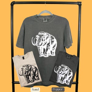 May include: Three t-shirts with a white silhouette of a mammoth on a dark grey, light beige, and dark grey background. The t-shirts are labeled "Sand", "Pepper", and "Pepper".