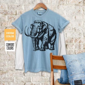 May include: A light blue t-shirt with a black and white illustration of a woolly mammoth. The shirt has a white underlay and is made of a soft, comfortable fabric.