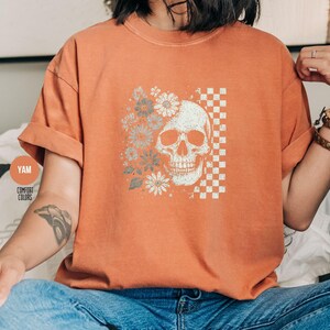 May include: Orange Comfort Colors t-shirt featuring a skull design with flowers and a checkered pattern. The skull is on the right side, with flowers on the left. The shirt has rolled-up sleeves.