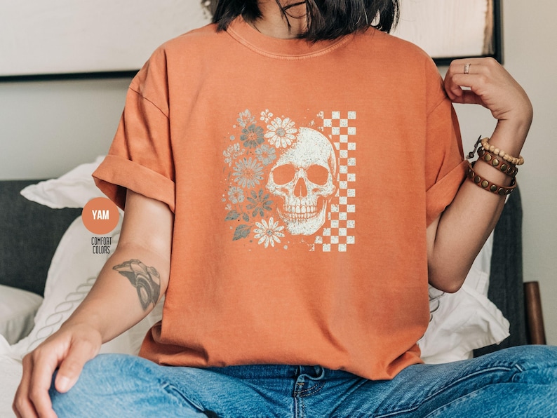 May include: Orange Comfort Colors t-shirt featuring a skull design with flowers and a checkered pattern. The skull is on the right side, with flowers on the left. The shirt has rolled-up sleeves.