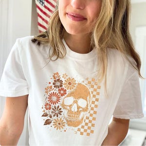 May include: White t-shirt featuring a skull design with floral and checkered elements in shades of brown and orange. The design is centered on the shirt. The shirt has a crew neck and short sleeves.