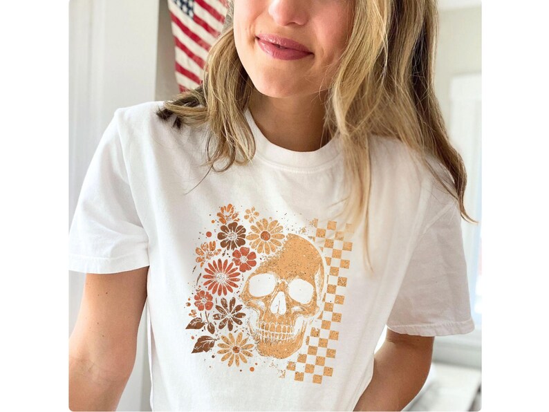 May include: White t-shirt featuring a skull design with floral and checkered elements in shades of brown and orange. The design is centered on the shirt. The shirt has a crew neck and short sleeves.