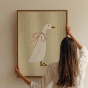 May include: A woman hangs a framed illustration of a white goose with a pink bow on a white wall. The illustration is on a striped background.