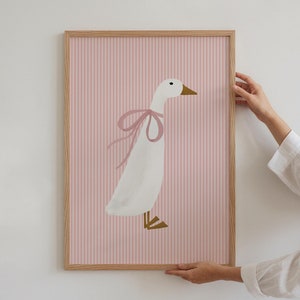 Könnte beinhalten: Ein gerahmter Druck einer weißen Gans mit einer rosa Schleife auf einem rosa-weiß gestreiften Hintergrund.