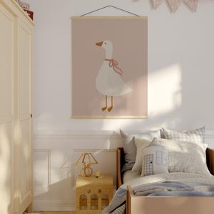May include: A children's room featuring a framed poster with a white goose illustration wearing a pink bow against a pink background. The room includes a bed with pillows, a wooden nightstand, and a decorative lamp.