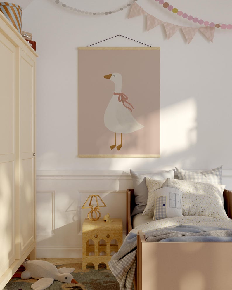 May include: A children's room featuring a framed poster with a white goose illustration wearing a pink bow against a pink background. The room includes a bed with pillows, a wooden nightstand, and a decorative lamp.