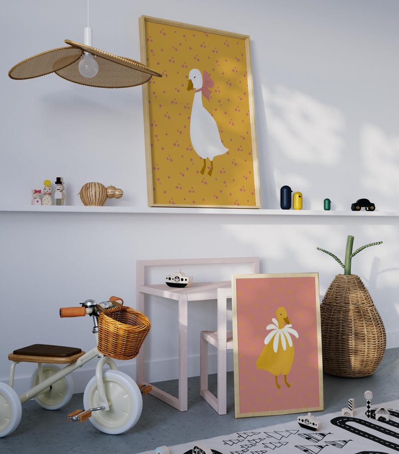 Pu&ograve; includere: Un'illustrazione di un'oca bianca e gialla con un fiocco rosa su uno sfondo giallo con puntini rosa. Una seconda illustrazione di un'oca gialla con un colletto di fiori bianchi su uno sfondo rosa. Entrambe le illustrazioni sono incorniciate in legno.