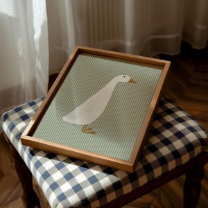 May include: A framed print of a white duck with a gold beak and legs on a green and white striped background. The frame is made of wood and has a natural finish.