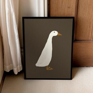 Pode incluir: Impress&atilde;o art&iacute;stica emoldurada com uma ilustra&ccedil;&atilde;o minimalista de um pato branco com bico e p&eacute;s amarelos. O pato est&aacute; sobre um fundo castanho com linhas verticais, dentro de uma moldura preta. A obra est&aacute; encostada a uma parede.