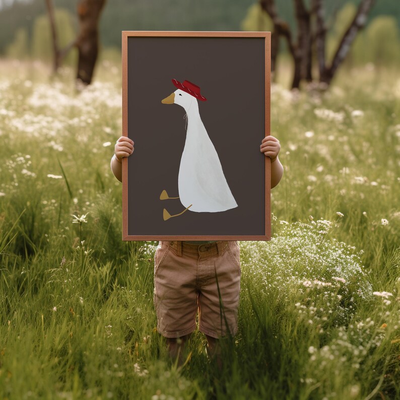 May include: A child holds up a framed illustration of a white goose wearing a red hat. The goose is sitting with its legs crossed and has a gold beak.