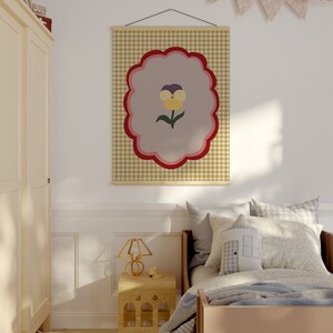 May include: A yellow, red, and purple flower with a green stem is printed on a beige and brown gingham fabric poster. The poster is hanging on a white wall in a bedroom with a bed, a nightstand, and a lamp.