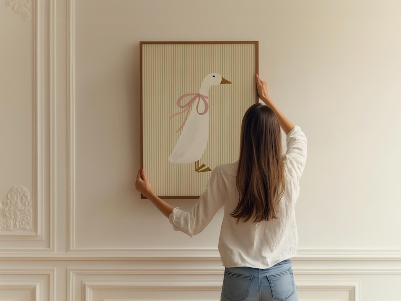 May include: A woman hangs a framed illustration of a white goose with a pink bow on a white wall. The illustration is on a striped background.