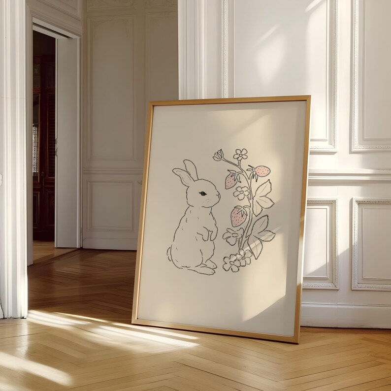May include: A framed illustration of a white rabbit standing next to a pink strawberry plant with green leaves and flowers. The background is a light beige color.