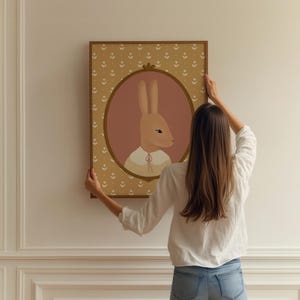 May include: A woman hangs a framed illustration of a brown rabbit with a pink background and a yellow floral pattern border. The rabbit is wearing a white collar with a yellow bow.