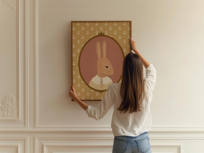 May include: A woman hangs a framed illustration of a brown rabbit with a pink background and a yellow floral pattern border. The rabbit is wearing a white collar with a yellow bow.