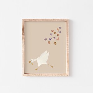May include: A framed print with a white goose running towards a flock of purple and orange butterflies on a beige background.