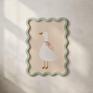 May include: A framed wall art print featuring a white goose with a pink ribbon around its neck. The goose is set against a light peach background, framed by a wavy, light green border. The artwork is mounted on a white wall.