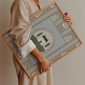 May include: A framed print with a light blue and white gingham pattern, a white house with green trees, and a floral border on a gray background.