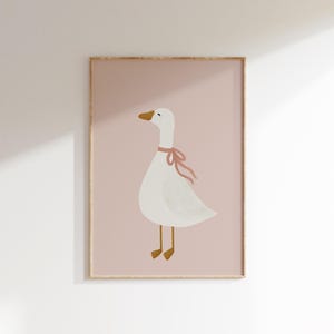 May include: A framed print featuring a whimsical illustration of a white goose with a pink ribbon around its neck. The goose has a yellow beak and feet, set against a soft pink background. The artwork is framed in a light wood tone.