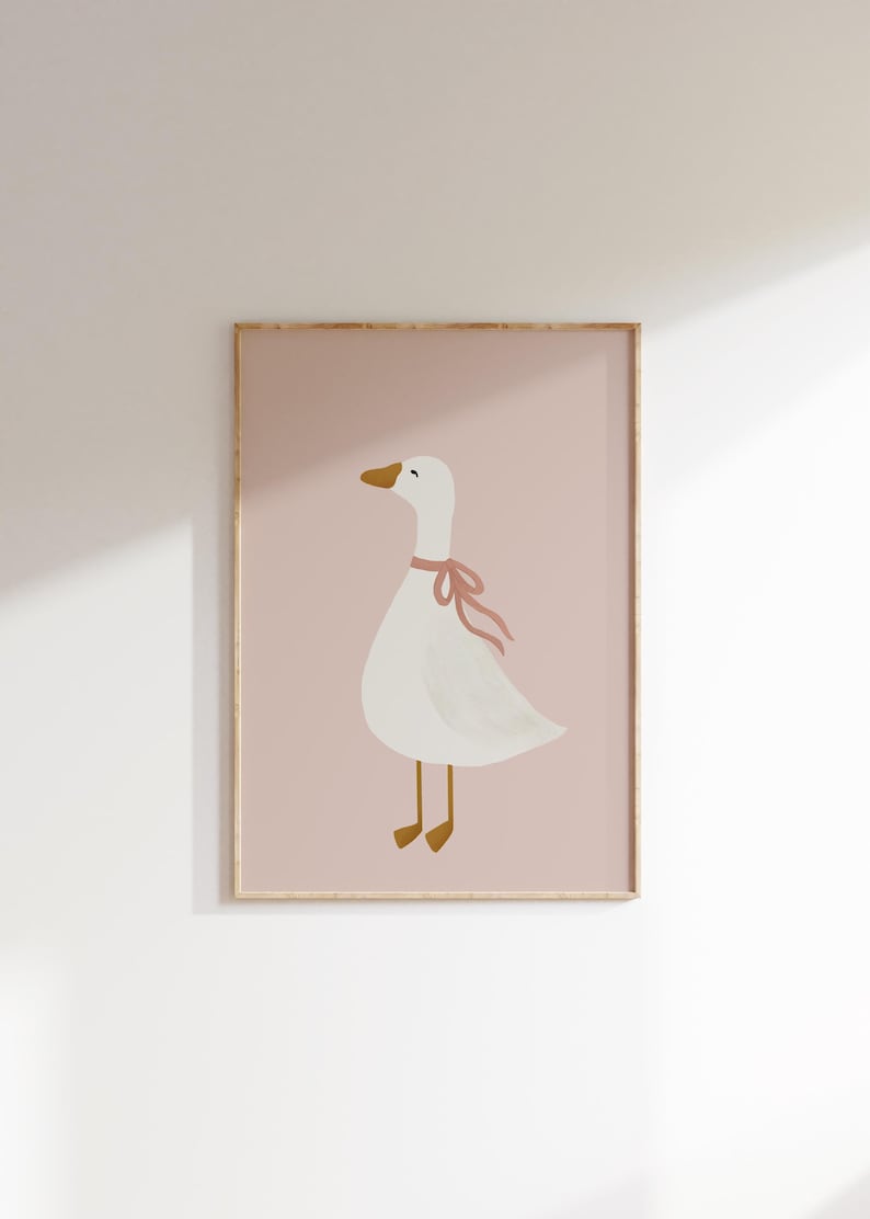 May include: A framed print featuring a whimsical illustration of a white goose with a pink ribbon around its neck. The goose has a yellow beak and feet, set against a soft pink background. The artwork is framed in a light wood tone.