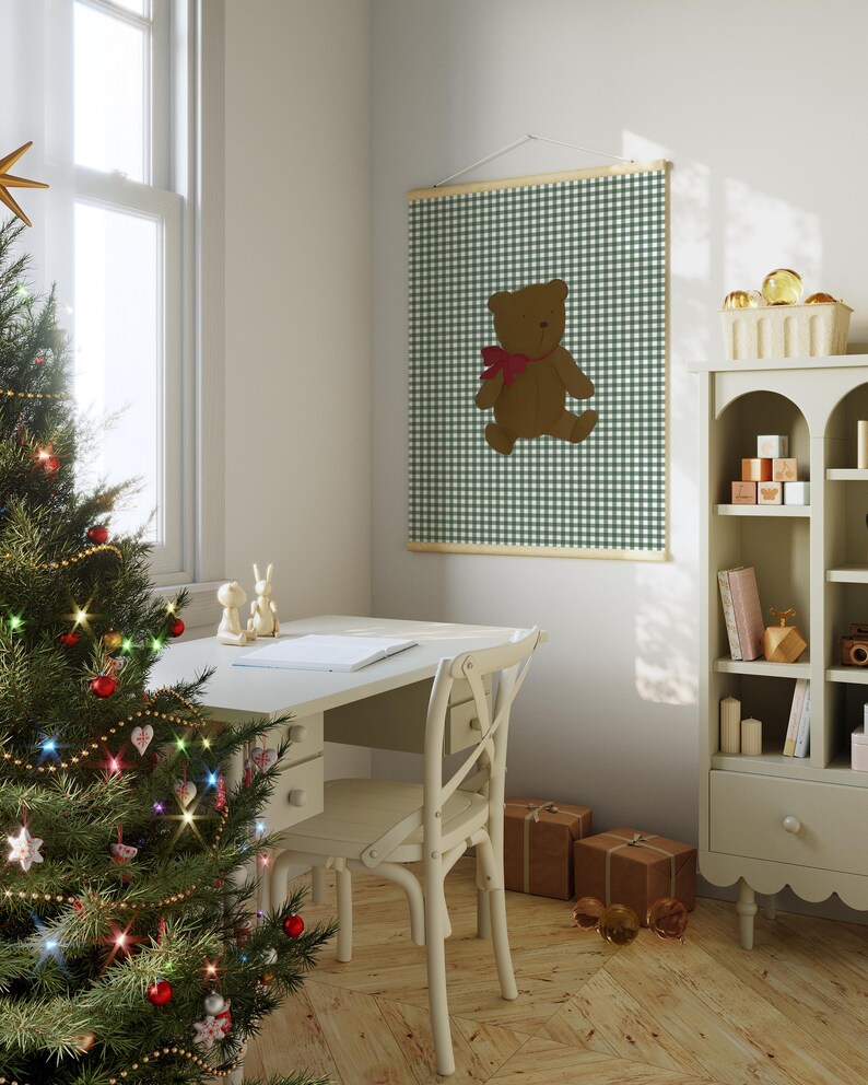 May include: A white wooden desk with a chair and a Christmas tree in the corner. A framed print of a brown teddy bear on a green and white gingham background hangs on the wall.