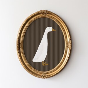 Pode incluir: Obra de arte oval com moldura dourada com uma ilustra&ccedil;&atilde;o de um pato branco sobre um fundo castanho listrado. O pato tem bico e p&eacute;s amarelos. A moldura tem detalhes ornamentais e est&aacute; colocada numa parede branca.