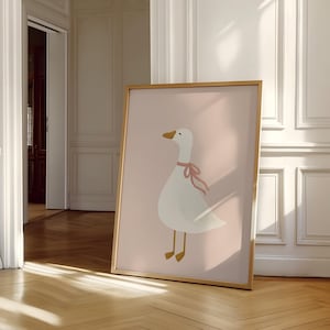 May include: A framed art print featuring a whimsical illustration of a white goose with a pink ribbon around its neck. The goose is set against a soft pink background. The frame is a warm gold color, adding a touch of elegance to the artwork.