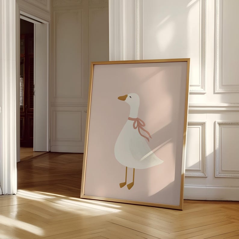 May include: A framed art print featuring a whimsical illustration of a white goose with a pink ribbon around its neck. The goose is set against a soft pink background. The frame is a warm gold color, adding a touch of elegance to the artwork.