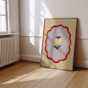 May include: A framed print with a yellow and white gingham pattern. The print features a pink, red, and white scalloped border with a purple and yellow flower inside.