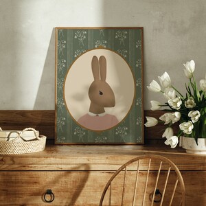 May include: A framed illustration of a brown rabbit wearing a pink shirt against a green background with a floral pattern. The frame is on a wooden table with a wicker basket and a vase of white tulips.