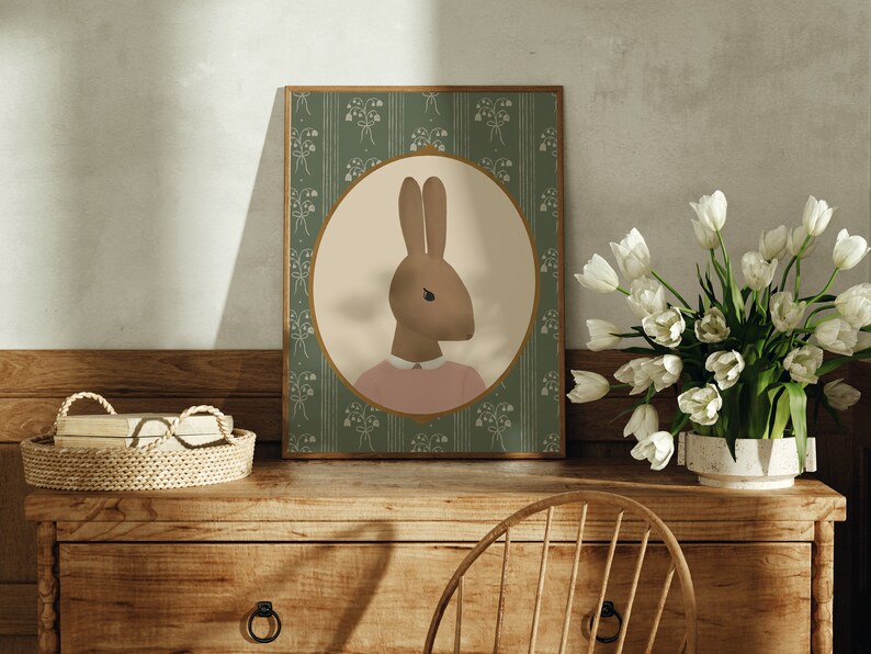 May include: A framed illustration of a brown rabbit wearing a pink shirt against a green background with a floral pattern. The frame is on a wooden table with a wicker basket and a vase of white tulips.