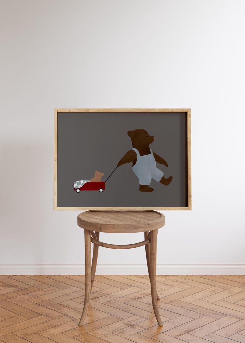 K&ouml;nnte beinhalten: Eine gerahmte Illustration eines braunen B&auml;ren, der eine Latzhose tr&auml;gt und einen roten Wagen mit einem kleineren braunen B&auml;ren darin zieht. Die Illustration ist auf grauem Hintergrund und wird auf einem Holzhocker ausgestellt.