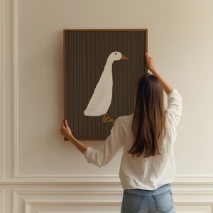 Pode incluir: Uma impress&atilde;o de arte emoldurada com uma ilustra&ccedil;&atilde;o de um pato branco sobre um fundo marrom escuro com listras verticais. A obra de arte tem uma moldura de madeira e est&aacute; sendo segurada contra uma parede branca.