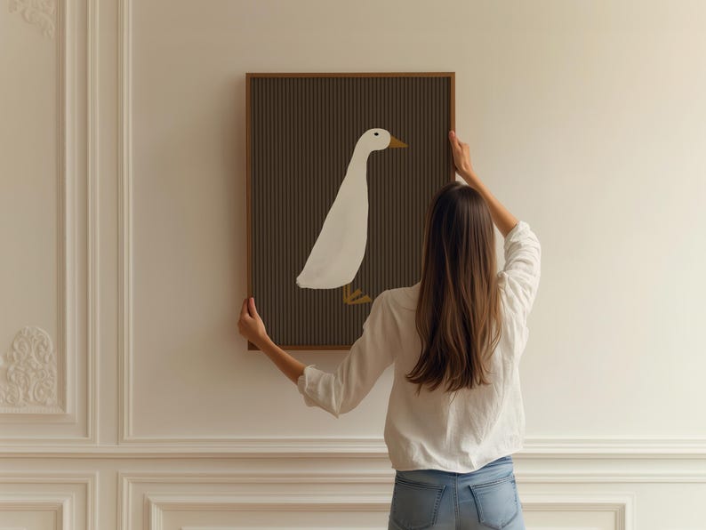 Pode incluir: Uma impress&atilde;o de arte emoldurada com uma ilustra&ccedil;&atilde;o de um pato branco sobre um fundo marrom escuro com listras verticais. A obra de arte tem uma moldura de madeira e est&aacute; sendo segurada contra uma parede branca.