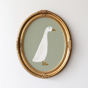 May include: A gold oval frame with a white duck illustration on a green and white striped background.