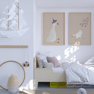 May include: A white wooden bed with a white duvet and pillows. There are two framed prints on the wall above the bed, one with a goose wearing a purple hat and the other with a goose flying with butterflies. There is a white wicker basket with a face on it on the floor next to the bed.