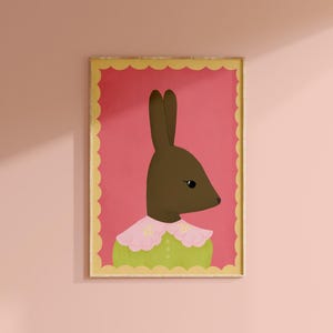 May include: A framed art print featuring a brown rabbit with a pink and green outfit. The rabbit has a pink collar with yellow flower accents. The background is a vibrant pink, and the frame has a scalloped edge.