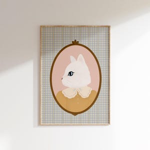 May include: A white cat with blue eyes wearing a yellow shirt with a lace collar is depicted in a pink oval frame with a gold border. The frame is set against a blue and white checkered background.