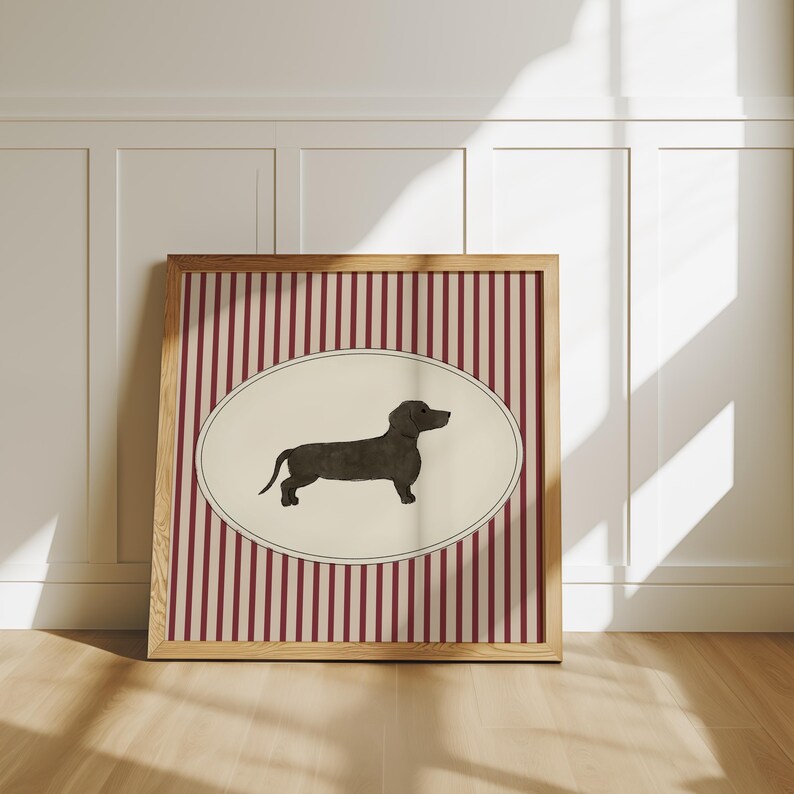 May include: A framed print featuring a dachshund illustration. The dog is depicted in a dark color against a cream-colored oval, set against a background of red and cream vertical stripes. The frame is made of light-colored wood.