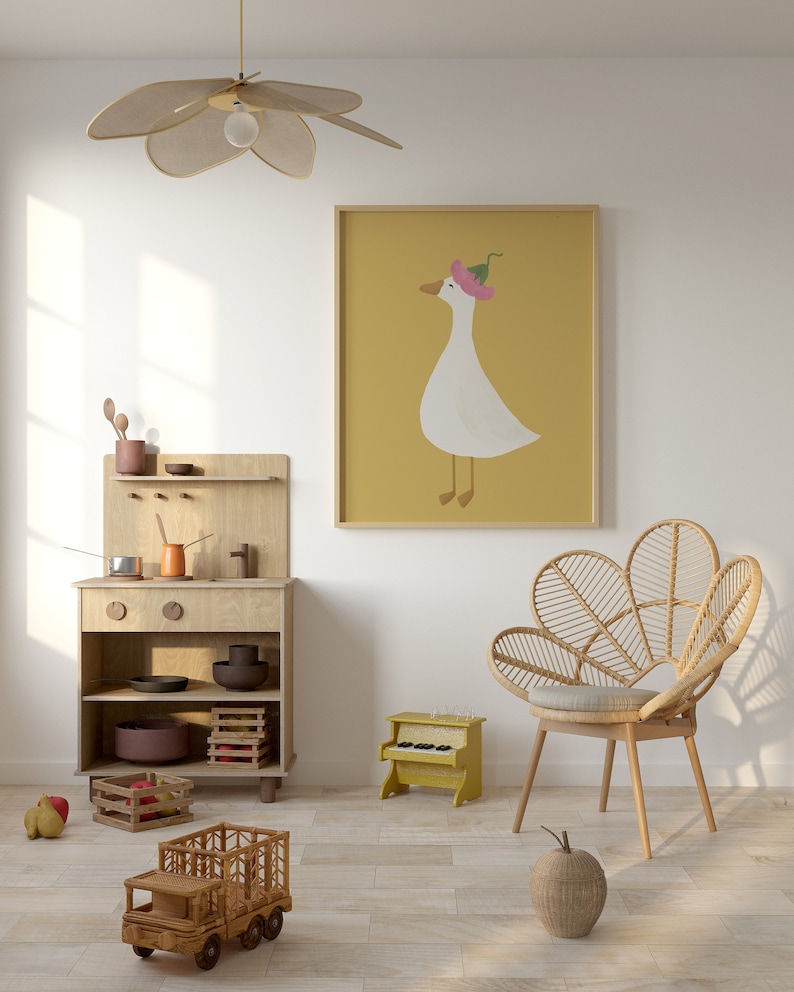 May include: A light wood play kitchen with a toy truck, a toy piano, and a wicker chair in a room with a white wall and a framed print of a goose wearing a pink flower on its head.