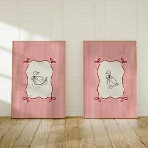 May include: Two pink framed prints with white backgrounds and red ribbon accents. Each print features a black and white line drawing of a duck with a ribbon around its neck.