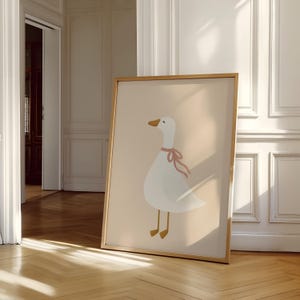 May include: A framed art print featuring a cartoon goose with a white body, a yellow beak and feet, and a pink ribbon around its neck. The goose is set against a light beige background. The frame is a light brown color.